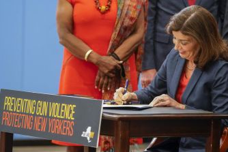 A woman sitting at a table signs legislation related to gun violence prevention, with a sign reading "PREVENTING GUN VIOLENCE PROTECTING NEW YORKERS" in front of her.