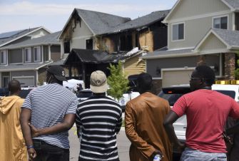 Group of people standing with their backs to the camera, watching a heavily burned suburban house with emergency responders nearby.