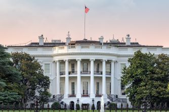 The White House exterior with an American flag flying atop, framed by trees under a pastel sunset sky.