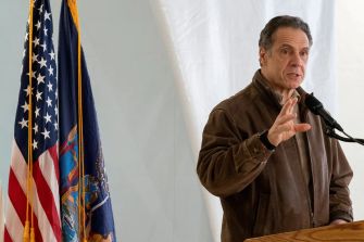 New York Governor Andrew Cuomo speaking at a press event, with the state flag in the background.