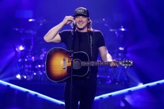 Morgan Wallen performing on stage with an acoustic guitar, wearing a black shirt and a cap.