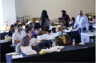 A group of people in an election office environment reviewing absentee ballots, with some individuals engaged in conversation and wearing masks.