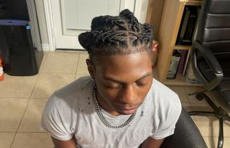 A Black teenager with twisted braids wearing a gray t-shirt and silver chain, sitting in a chair with eyes closed.